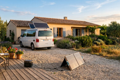 Camping-car équipé de panneaux solaires devant une maison avec un kit solaire à brancher posé au sol